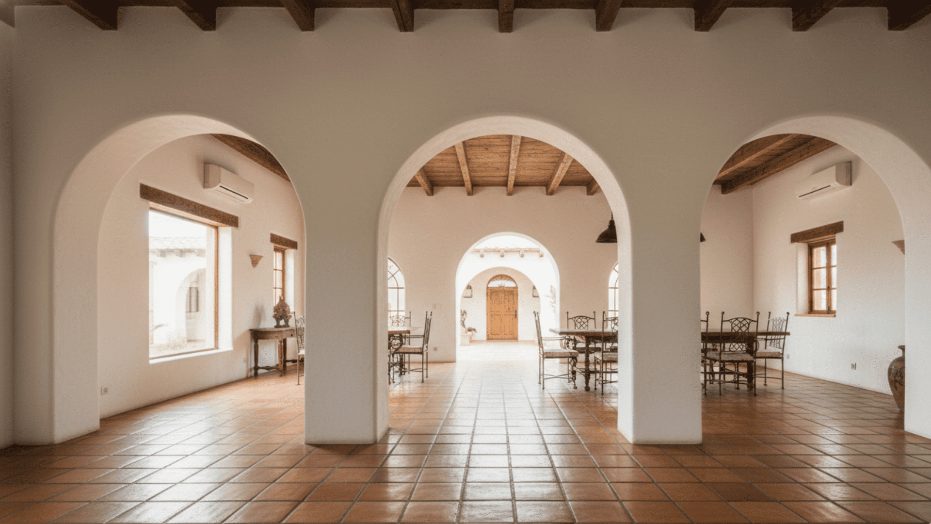 A hallway featuring white plastered arched doorways leading into a sunlit room with tile floors.