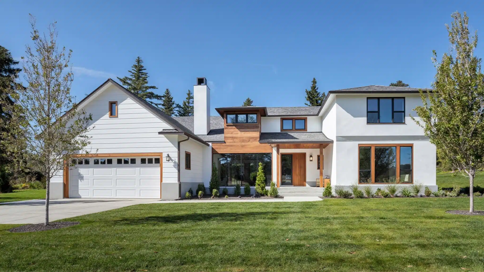 A modern two-story farmhouse with white siding, large windows, and a wooden accent entryway under a clear blue sky.