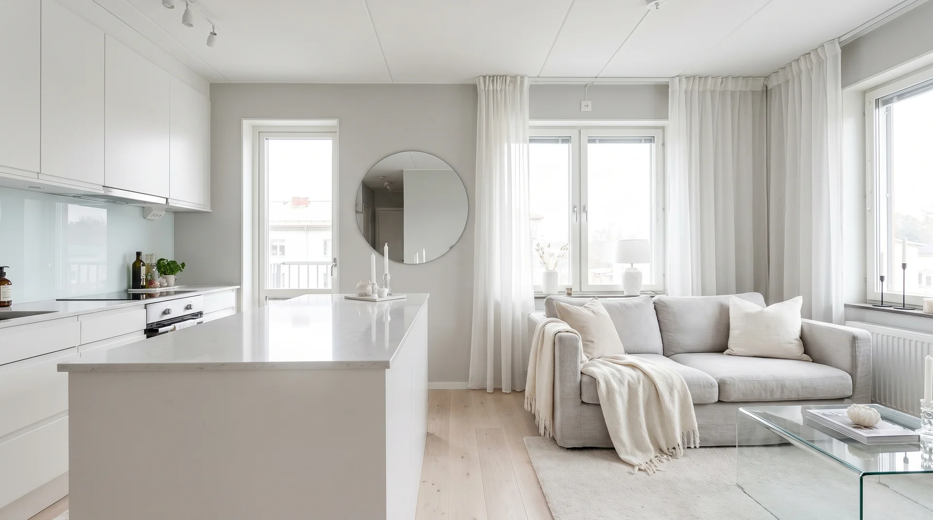 Bright all-white open-concept kitchen and living room featuring an island, a light grey sofa, and a round wall mirror.