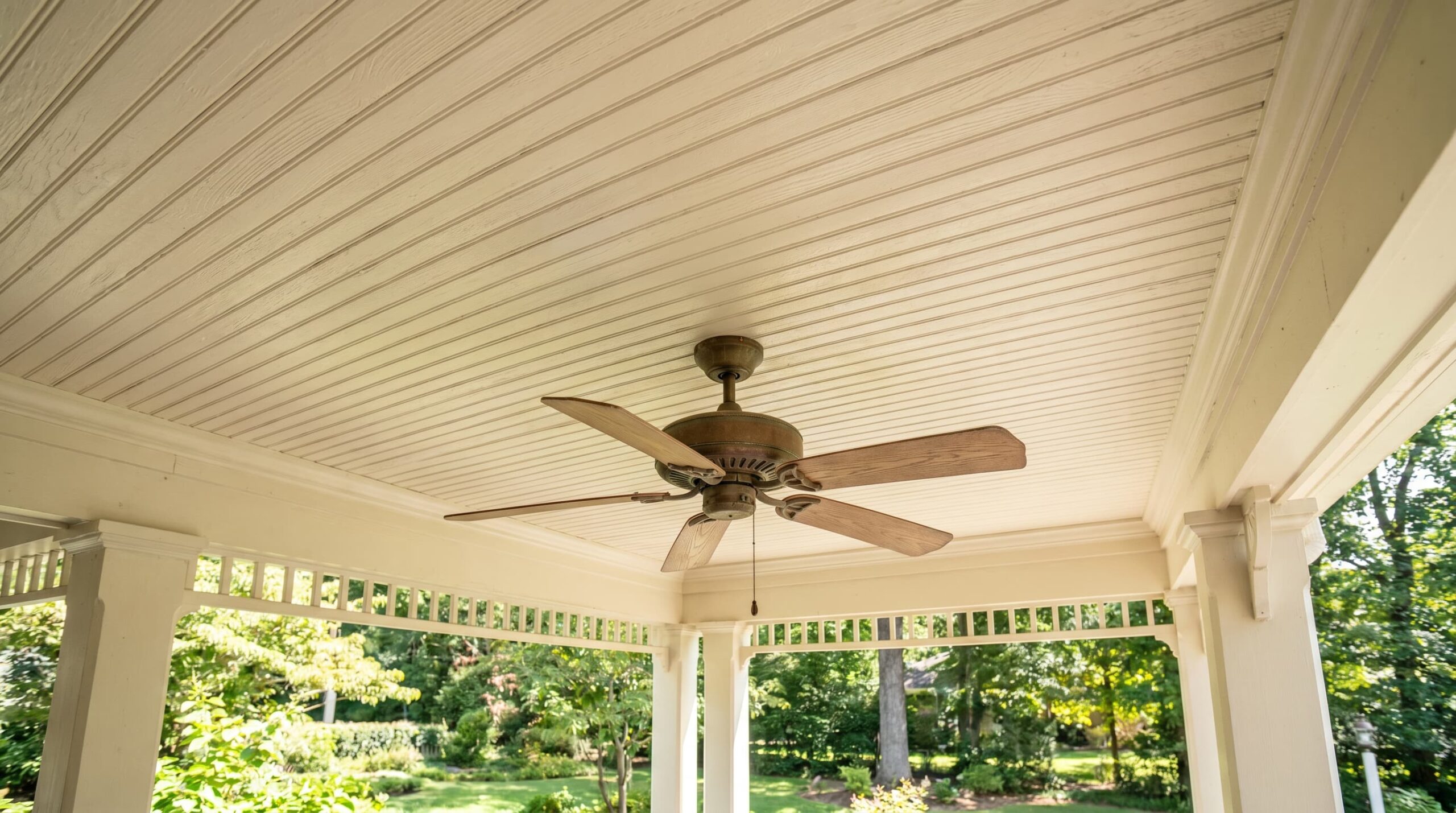 White covered porch ceiling with wooden beams, ceiling fan, and green garden view.