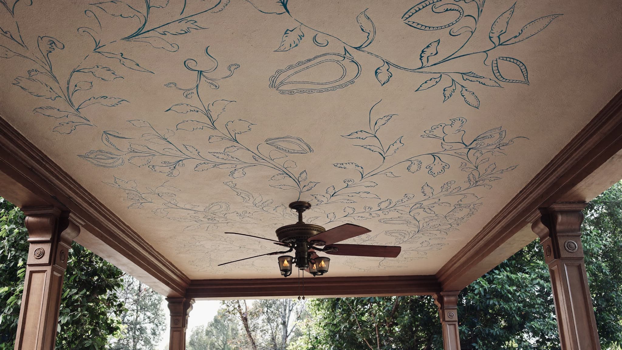 Covered porch with decorative floral ceiling, ceiling fan, wooden support columns, and garden view.