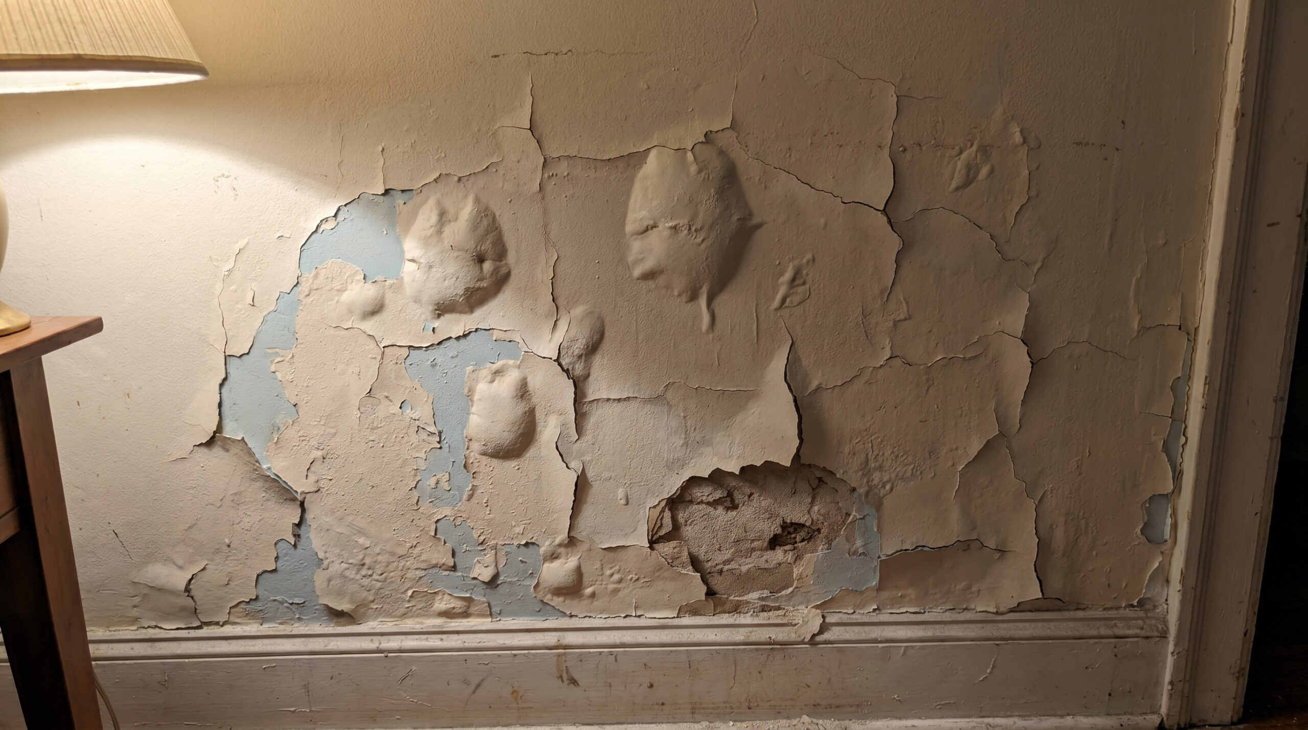 Close-up of severely cracked, bubbling, and peeling beige paint on an interior wall near baseboard trim.