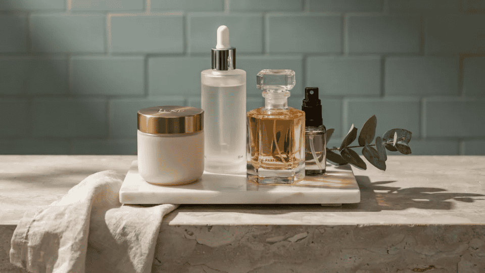Marble tray on bathroom shelf with skincare bottles, perfume, and a sprig of greenery in a spa-inspired setting