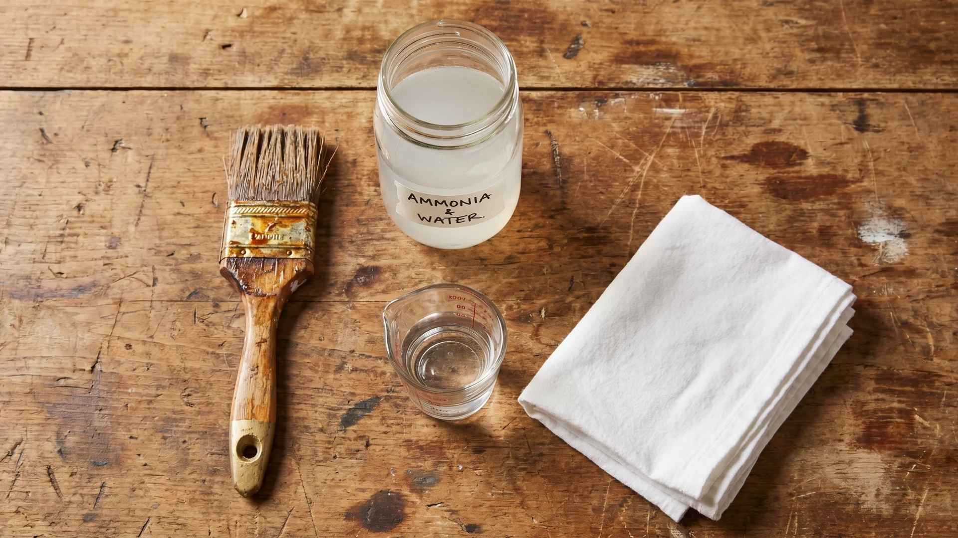 Paintbrush, ammonia solution, cloth, and measuring cup displayed on a rustic wooden table.