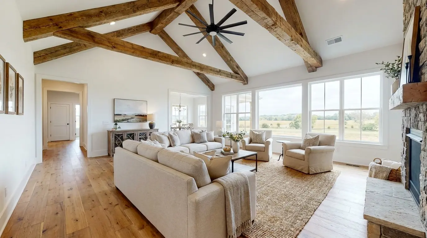 Modern farmhouse living room with exposed wooden ceiling beams, cream seating, large windows, and a stone fireplace.